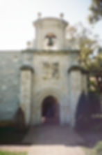 Stone archway of a historic building with detailed carvings and a bell. Sunlight filters through trees, casting shadows on the path.