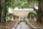 Outdoor wedding setup with wooden chairs and floral arrangements under string lights. Trees and a yellow building are in the background.