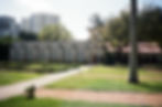 Historic stone wall with arched entrance in a garden setting. Tall buildings loom in the hazy background, with lush green trees nearby.
