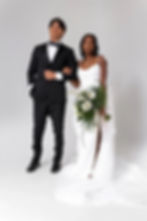 Bride in white dress holding bouquet stands arm-in-arm with groom in black tuxedo, set against a plain white background.