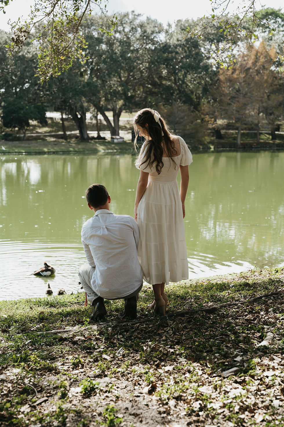 women standing in white dress and men kneeling beside her gaze out at the water while looking at the ducks 