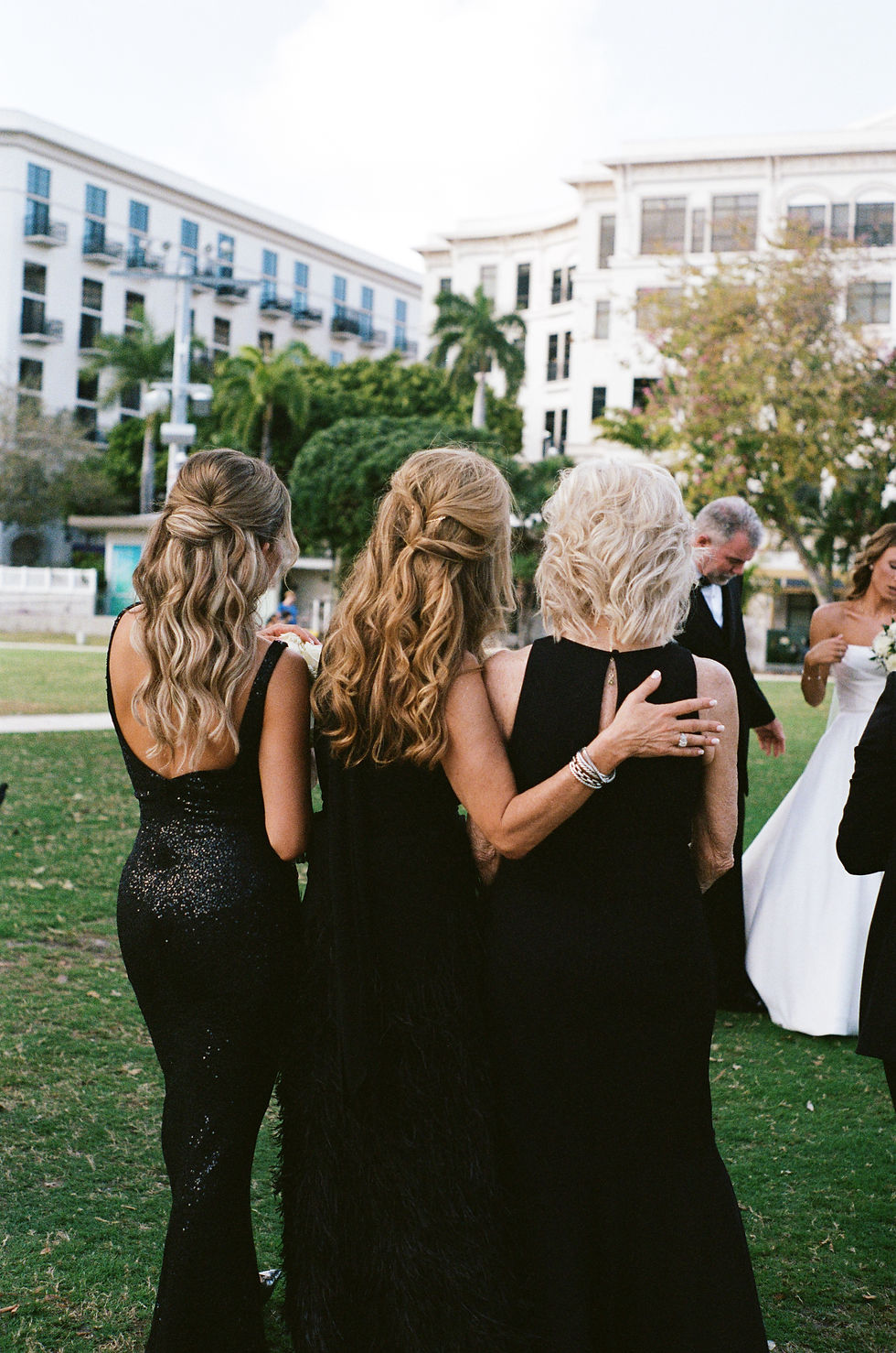 Three women in elegant black dresses stand arm-in-arm on grass, facing a modern building, conveying a warm, celebratory mood.