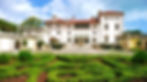 Elegant mansion with red roof and arched windows, surrounded by lush green gardens and manicured hedges under a blue sky.