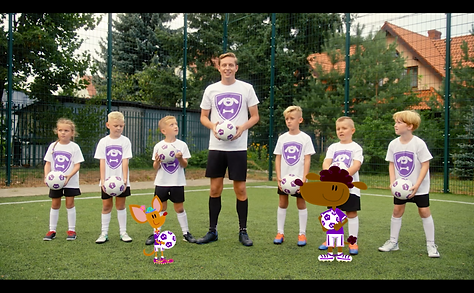 Psiaki Futbolaki Still: Michał the coach, 6 children and 2 animated Footypups stand and smile for the camera while holding footballs.