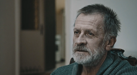 Caregiver Film Still: Yacek sits, his face is tired and drawn. He has grey hair and a white beard, he wears a grey sweater.