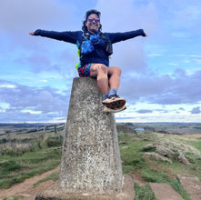 Woman sat on top of a stone trig point with arms outstretched.