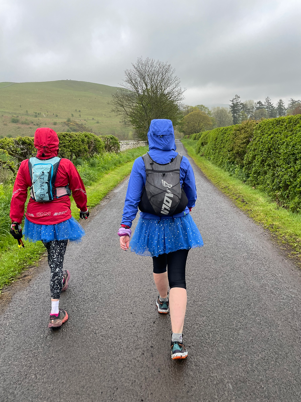 Two people in colorful jackets walk on a rural road, wearing blue tutus. Overcast sky, lush green hills, and hedges in the background.