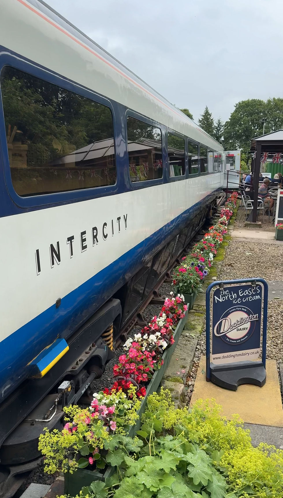 Train car labeled "INTERCITY" beside colorful flowerbeds. Sign reads "The North East's Ice Cream" by Doddington Dairy. Outdoor setting.