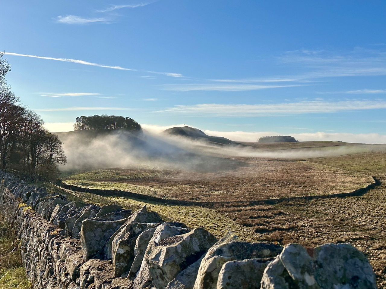 Stone wall in a field with fog over the hills under a blue sky.