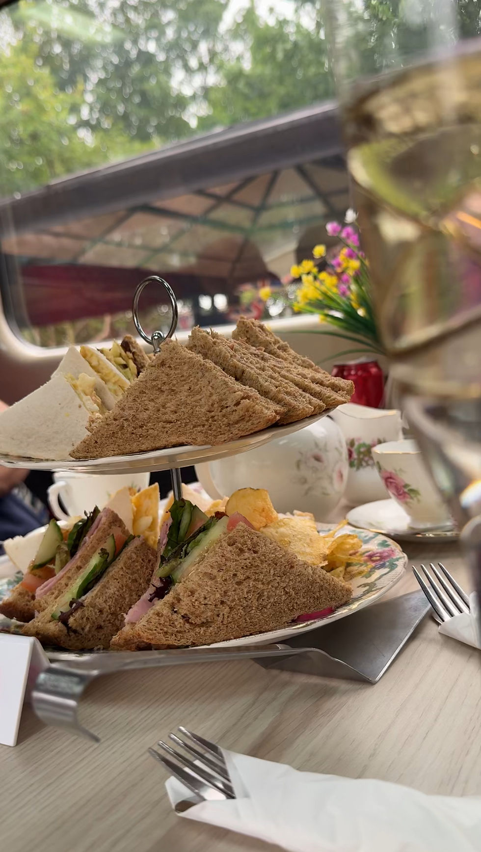Afternoon tea with tiered sandwich platter, teacups, and flowers on a table. Greenery visible outside, creating a cozy setting.