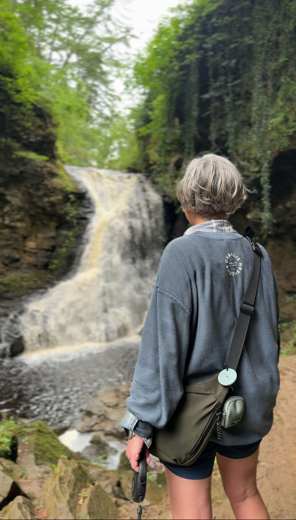 Person with short gray hair stands by a waterfall, holding a leash. They're wearing a blue sweater with a bag. Lush greenery surrounds them.