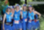 Five women in blue running outfits and tutus pose joyfully outdoors, showing peace signs. Their shirts read "It's not about the pace".