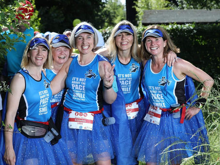 Five women in blue running outfits and tutus pose joyfully outdoors, showing peace signs. Their shirts read "It's not about the pace".