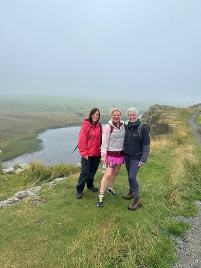 Three women smiling on a green hill with lake, Hadrian's Hike for Tynedale Hospice.