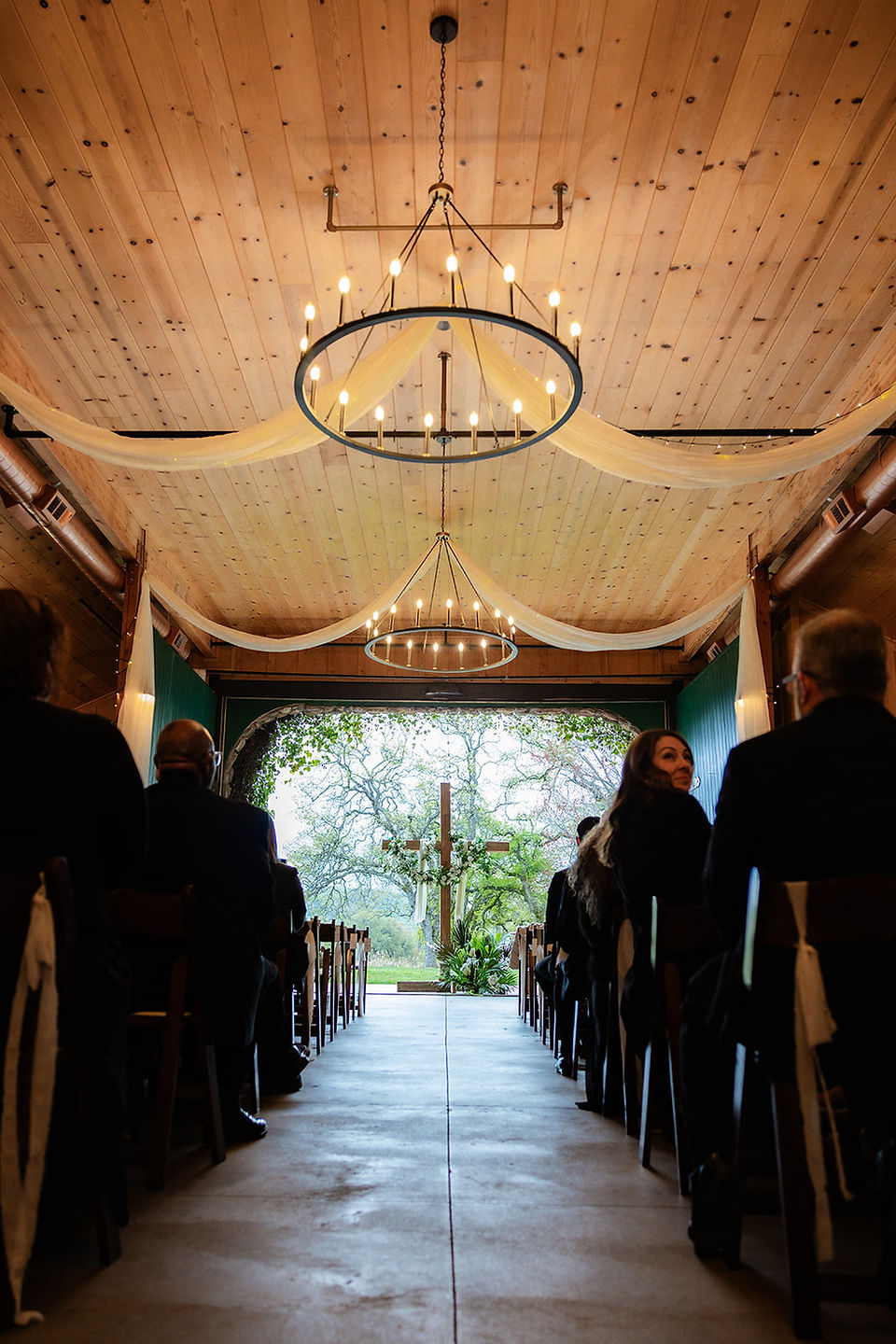 Indoor wedding ceremony at River Highlands Ranch with wooden cross altar, draped fabric, chandeliers, and guests seated