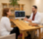 Médico conversa com paciente em consultório acolhedor, com monitor exibindo gráficos coloridos ao fundo.