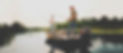 Two men fishing on a boat in a calm lake, surrounded by trees and greenery, under a clear sky.
