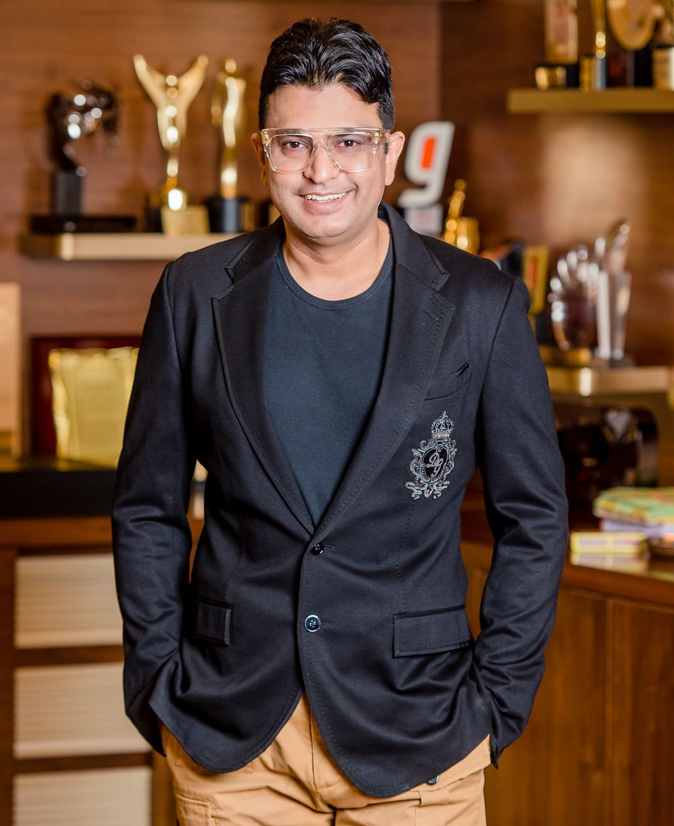 Man in a black blazer and tan pants smiling, with hands in pockets. Awards and trophies on shelves in a warmly lit background.