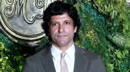 A man in a suit and tie stands in front of a lush green wall with a decorative emblem. The setting appears formal and elegant.