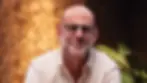 Bald man with glasses smiles softly against a warm, textured background with green leaves. Wears a light shirt, conveying a calm mood.