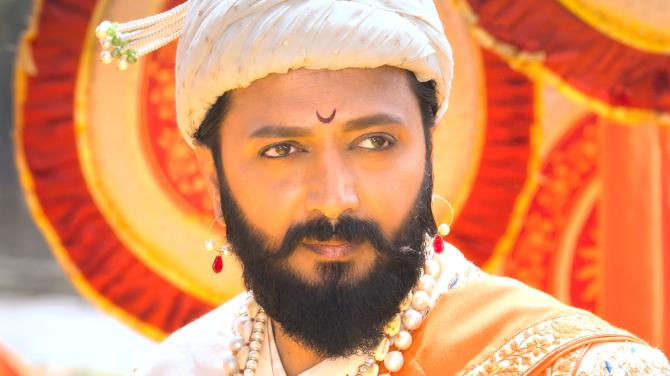 Man in traditional attire with a turban and pearl necklace, gazing thoughtfully. Vibrant background with orange and red patterns. Mood is regal.