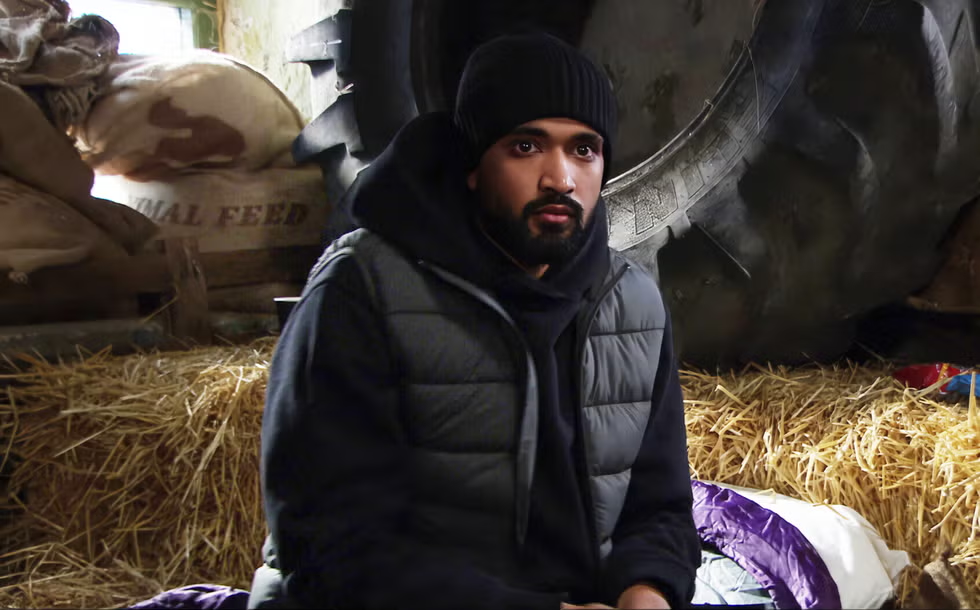 Man in black jacket and beanie sits on hay in a barn. Large tire and sacks labeled "animal feed" in the background. Calm expression.