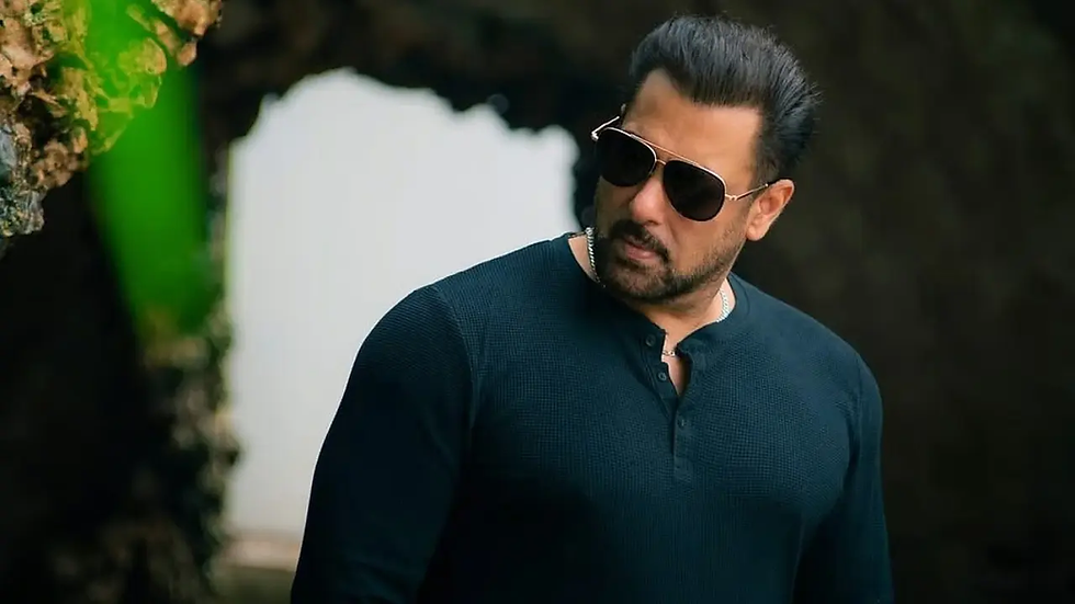 Man in sunglasses and dark shirt stands outdoors near a rocky arch and greenery, looking left with a serious expression.
