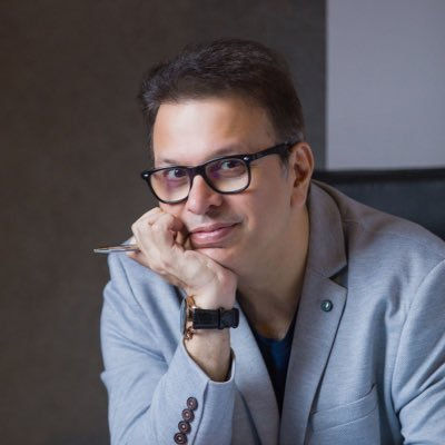 Man in glasses and gray blazer smiling, resting chin on hand with a pen. Neutral background, relaxed atmosphere.