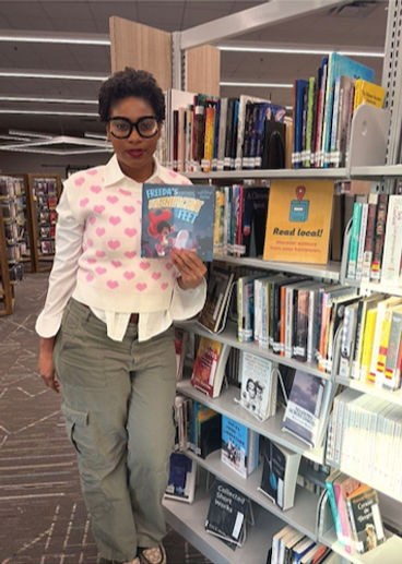 Kiyoko M. Hairston holding Freeda’s Fabnificent Feet beside library bookshelves.