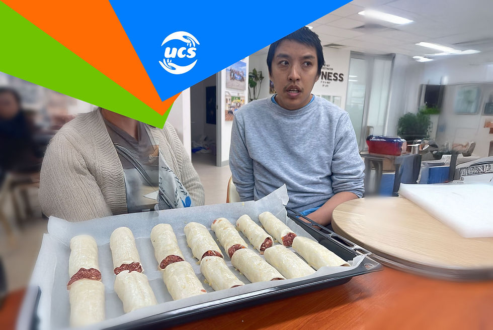 N. seated behind a tray of uncooked sausage rolls lined up on baking paper. Another person is partially visible to the left.