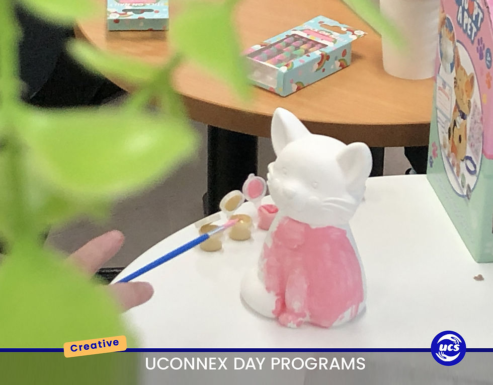A close-up of a white ceramic cat statue on a white table, with its lower body being painted pink. A hand holding a blue-handled paintbrush is visible on the left, dipping into a small pot of pink paint. Several other small pots of paint (gold and light pink) are also on the table