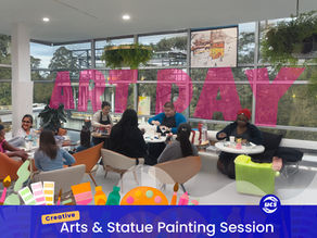 A group of people sitting at tables in a bright room with large windows, engaged in an arts and crafts activity.