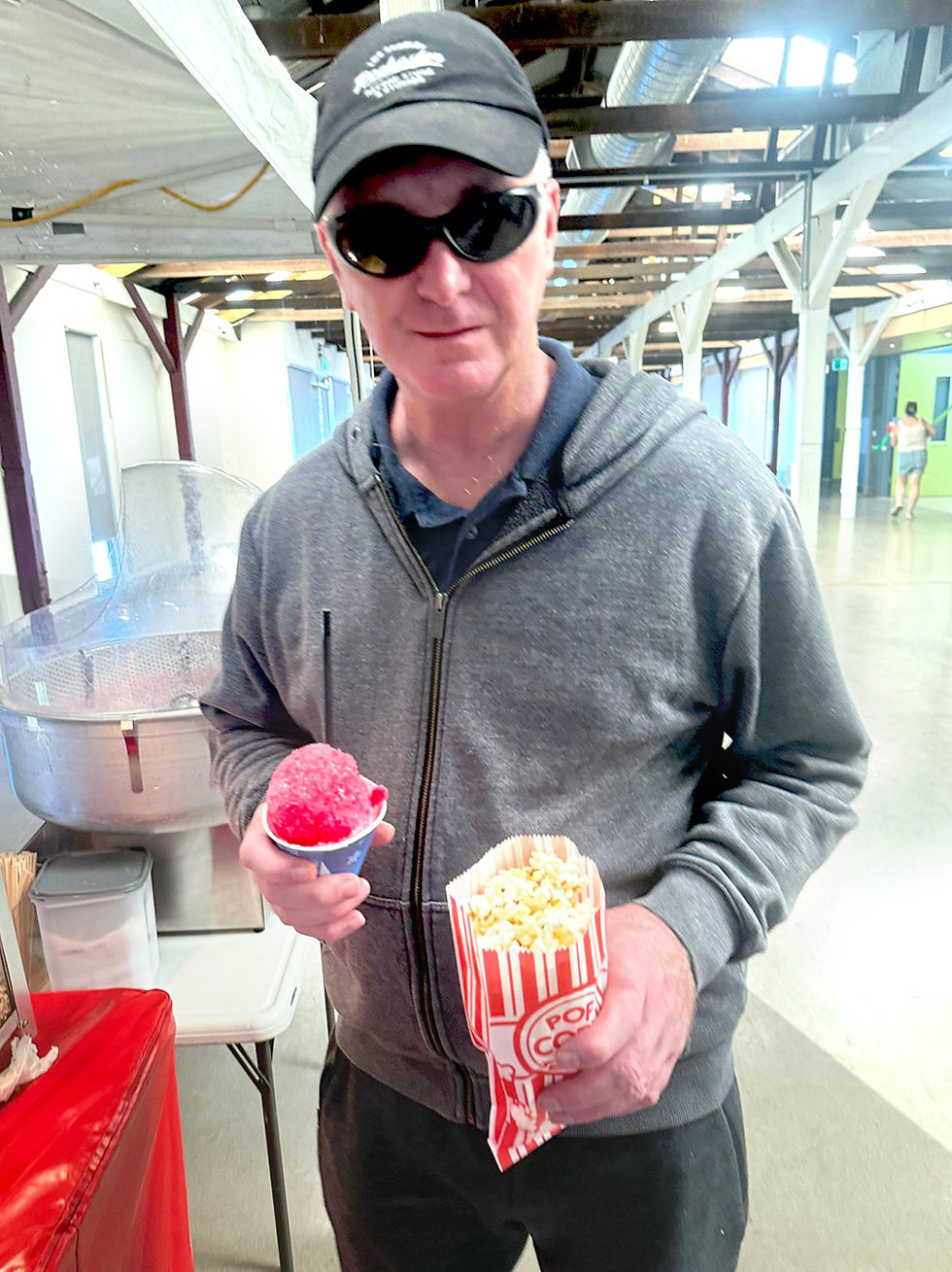 A man in a grey zip-up hoodie and sunglasses holds a red snow cone and a striped bag of fresh popcorn at the Shepparton Easter Festival.