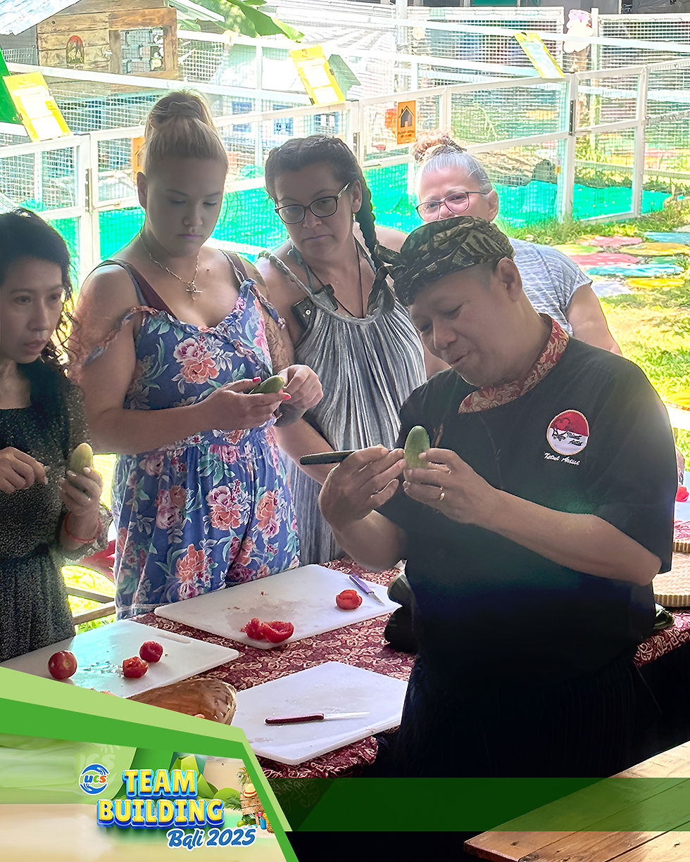 A group of Uconnex team members in Bali watches closely as a local chef, wearing a traditional Balinese headscarf, demonstrates intricate food carving techniques on a small green vegetable during the team building cooking challenge.