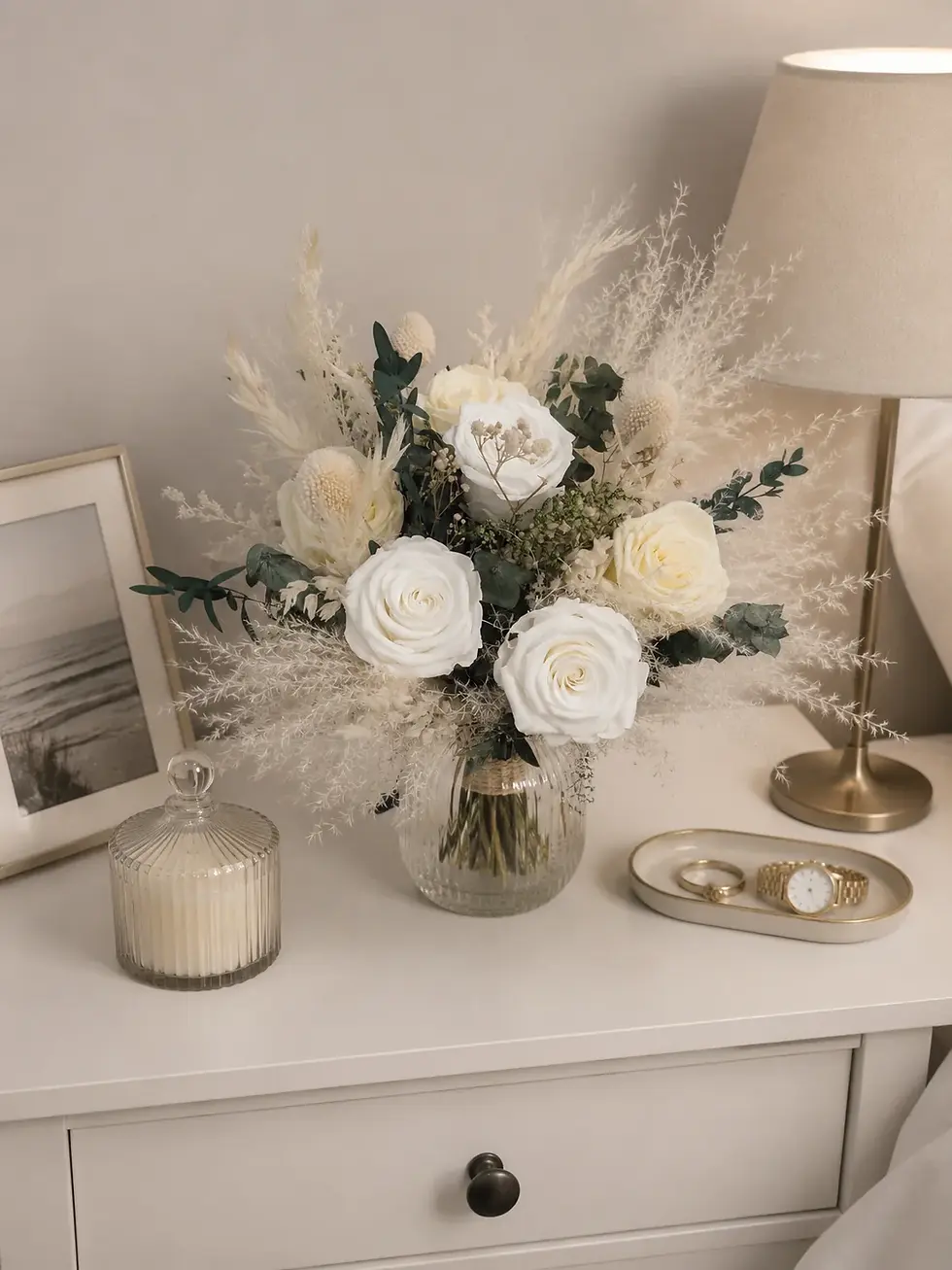 Luna Bloom ivory rose wedding bouquet with eucalyptus and cream foliage in a glass vase on a bedside table.