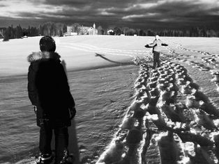 A black and white photo of two figures, one with arms outstretched in a field of snow with a church in the background