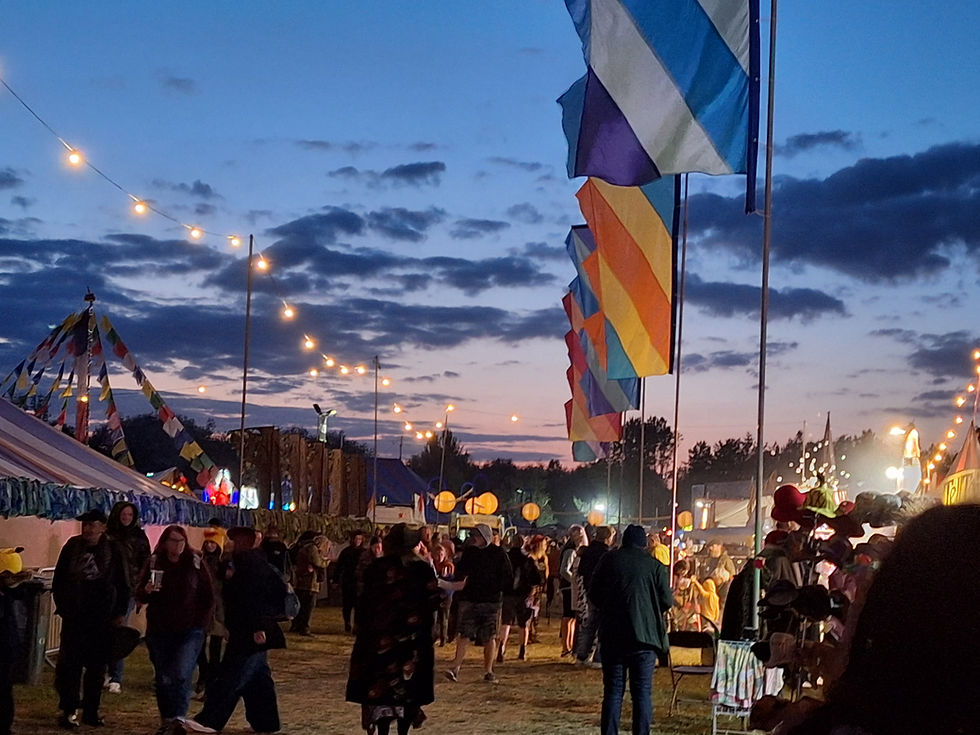 People moving through an outdoor festival marketplace at dusk, with illuminated stalls and colourful flags at Bearded Theory Festival.