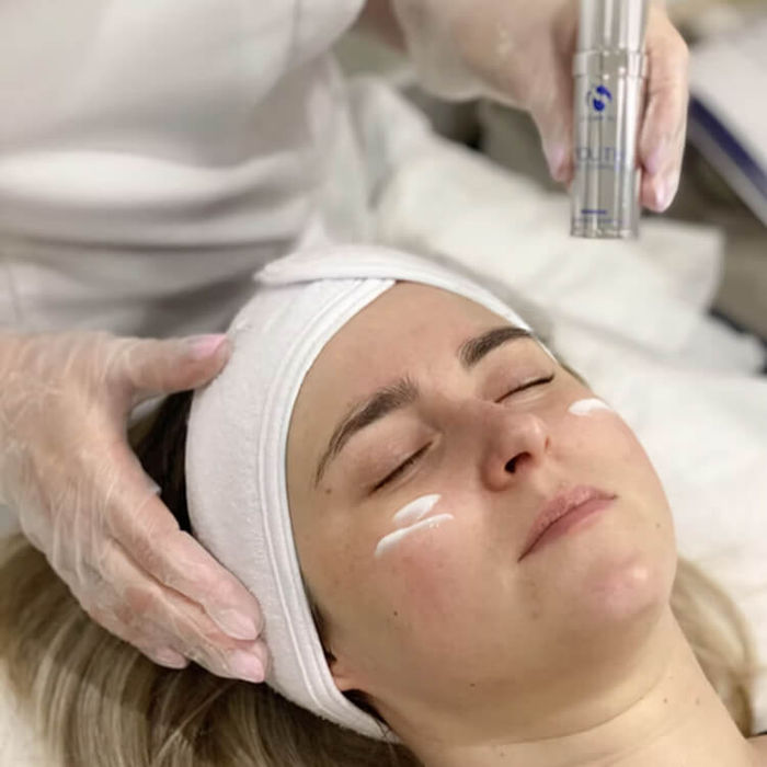 Aesthetician applying iS Clinical uk skincare product to a woman's face during a professional facial treatment, with visible white cream under the eyes