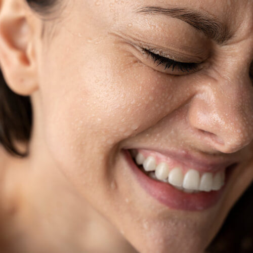 Close-up of a smiling woman’s face covered in water droplets, showcasing dewy, radiant skin — symbolizing the refreshing effects of the Hydration IV Booster