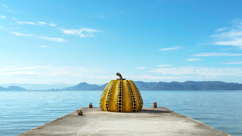 Yellow pumpkin sculpture with black polka dots on a pier, surrounded by calm blue sea, distant mountains, and a clear sky.