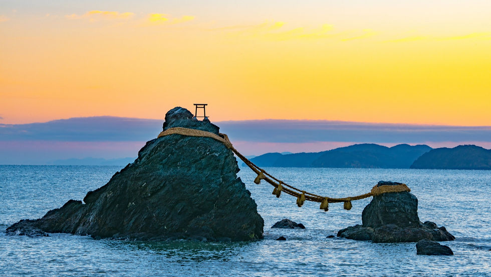 Two rocks connected by a rope in the ocean at sunrise. A small torii sits atop one rock. The sky displays hues of orange and yellow.