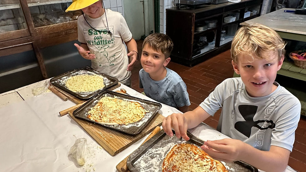 Three kids make pizzas at a kitchen table, smiling. One wears a yellow hat and a “Save our Wildlife” shirt. Brown tiled floor visible.