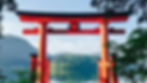 Red torii gate with black accents in serene lakeside setting, lush green hills in background, calm water reflects the blue sky.