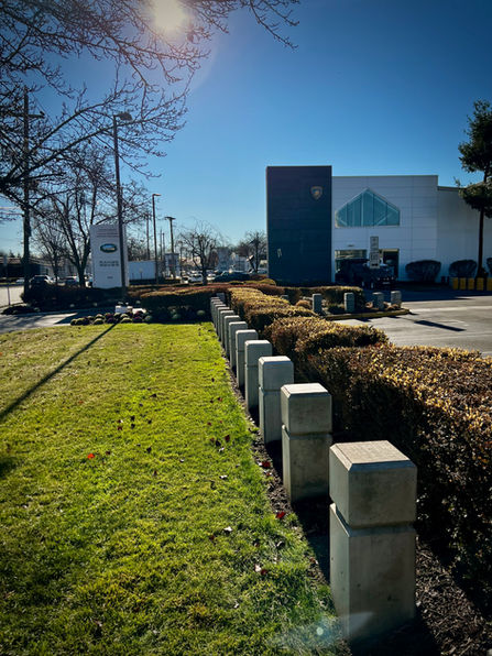 Line of custom concrete bollards along a landscaped pathway at a Lamborghini dealership in New Jersey, with hedges and a grassy lawn under a clear blue sky.