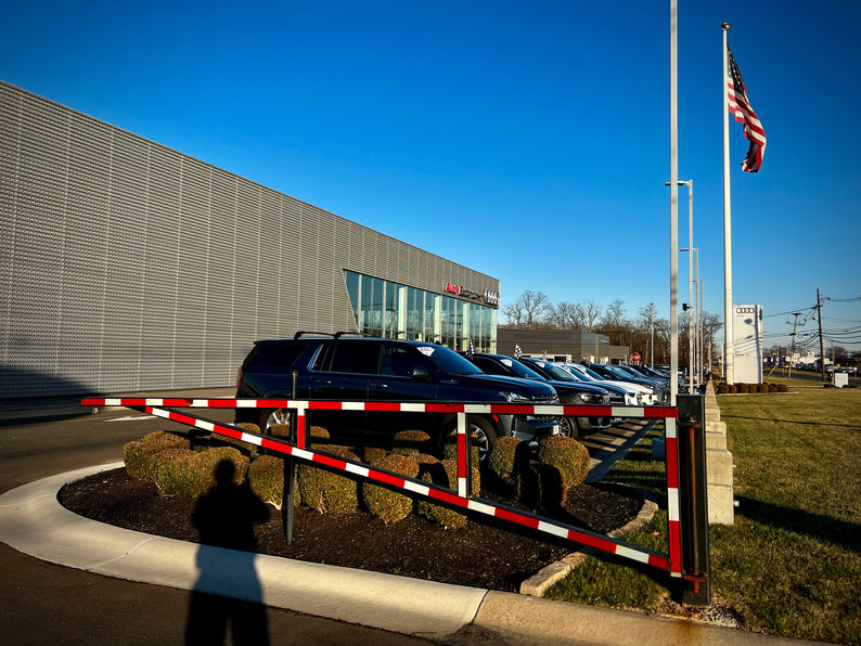 Photo of the Audi of Eatontown dealership in New Jersey with custom security crash gates in the foreground, luxury cars on display, an American flagpole, and a modern building exterior.