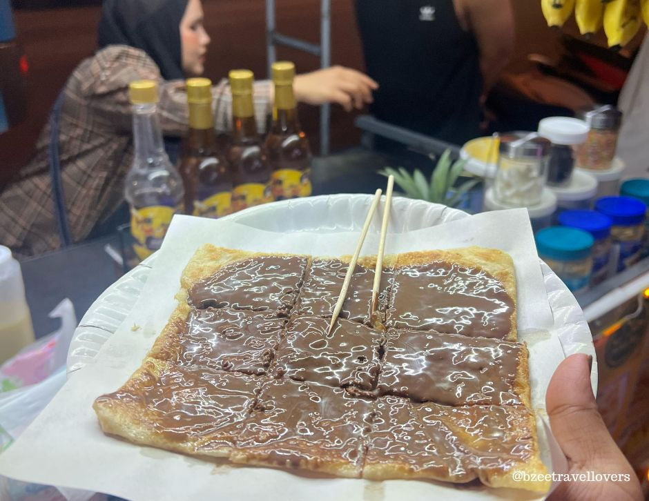 A close-up of a Nutella-covered roti on a paper plate with bamboo skewers. Background shows bottles and a woman reaching towards them. Thailand travel from India.