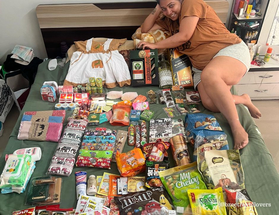 Woman smiling, lying beside a bed filled with various colorful food items, drinks, gifts, clothes, and a toy. Cozy, cheerful setting. Thailand travel from India.