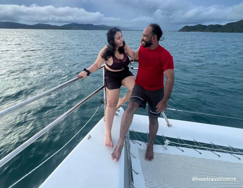 A couple smiles on a yacht with an ocean backdrop. The woman in a black outfit lounges on the railing, while the man in a red shirt sits. Thailand travel from India.