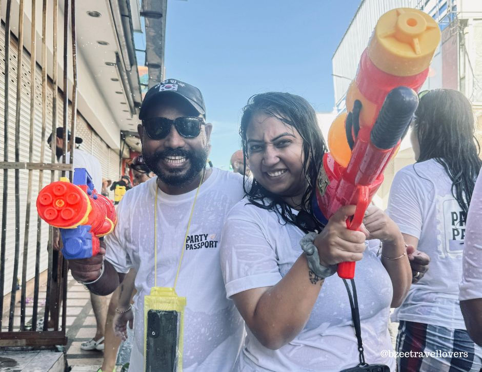 Two people smile holding colorful water guns in a street. They wear wet white shirts with "Party Passport." Bright, sunny day. Thailand travel from India.