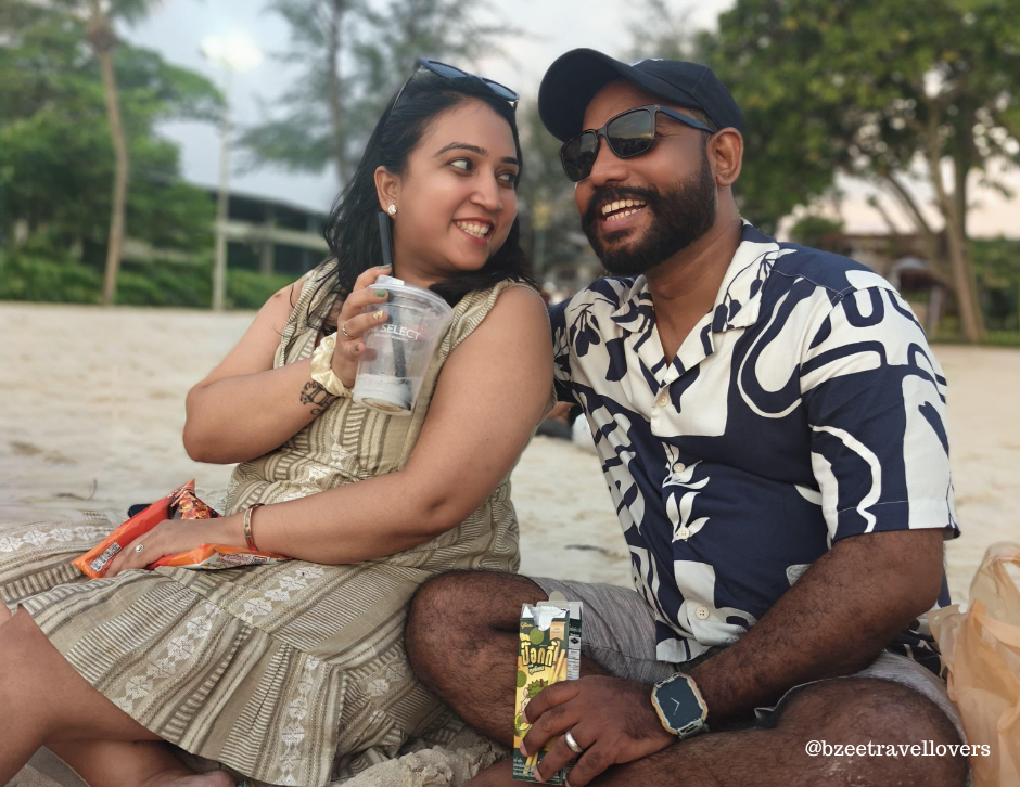 Smiling couple sitting on a beach, holding drinks and snacks. Green trees and greenery in the background. Casual summer attire. Sand visible.